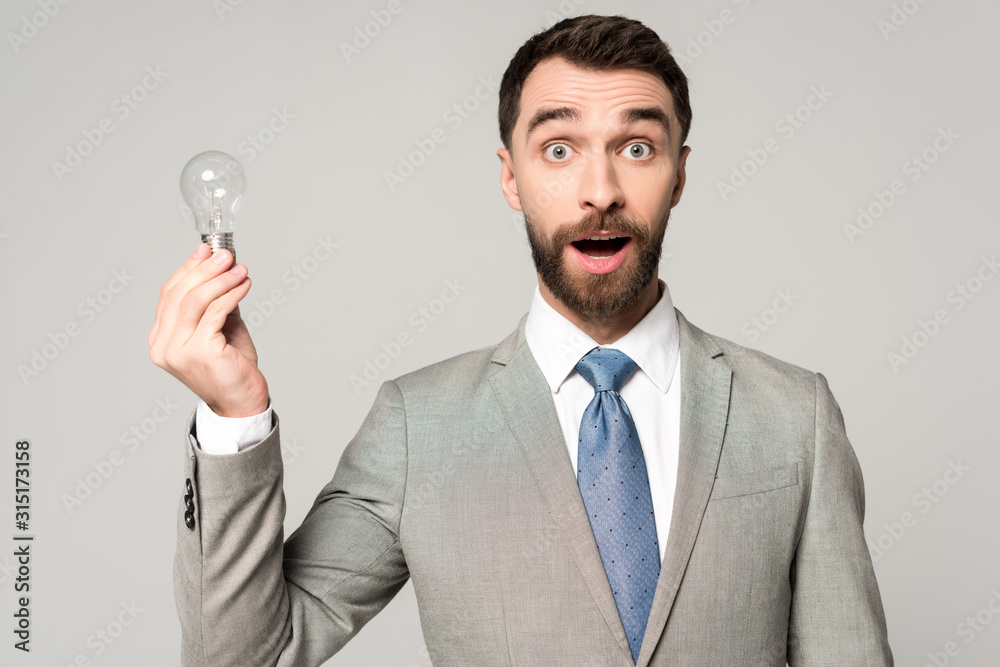 excited businessman holding light bulb and looking at camera isolated on grey excited businessman holding light bulb and looking at camera isolated on grey