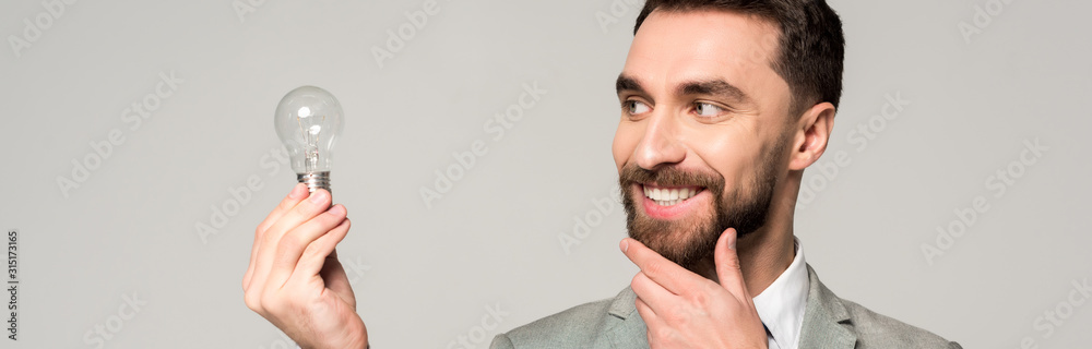 Obraz premium panoramic shot of smiling businessman touching face while holding light bulb isolated on grey