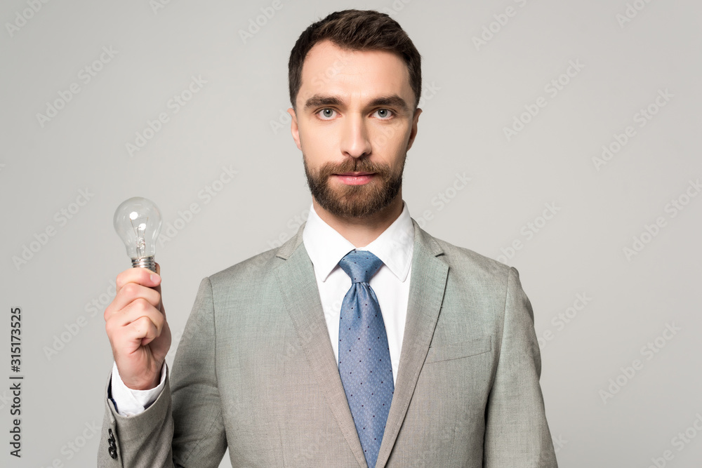 confident businessman holding light bulb and looking at camera isolated on grey confident businessman holding light bulb and looking at camera isolated on grey