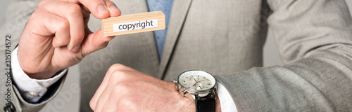 cropped view of businessman in wristwatch holding wooden block with copyright lettering isolated on grey, panoramic shot