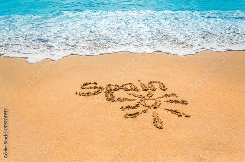 Hand written text Spain and sun symbol on the golden beach sand with coming wave