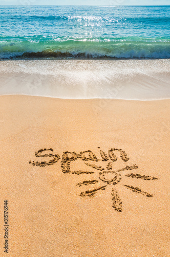 Hand written text Spain and sun symbol on the golden beach sand with coming wave. Vertical