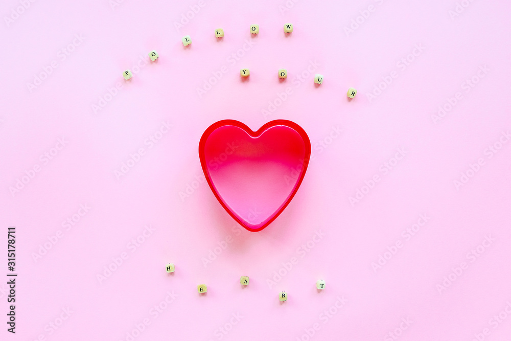 Creative holiday concept of love. Cookie cutter in a shape of heart on a pink background. Text Follow Your Heart made of wooden beads.  Valentine's Day concept. Top view, flat lay.