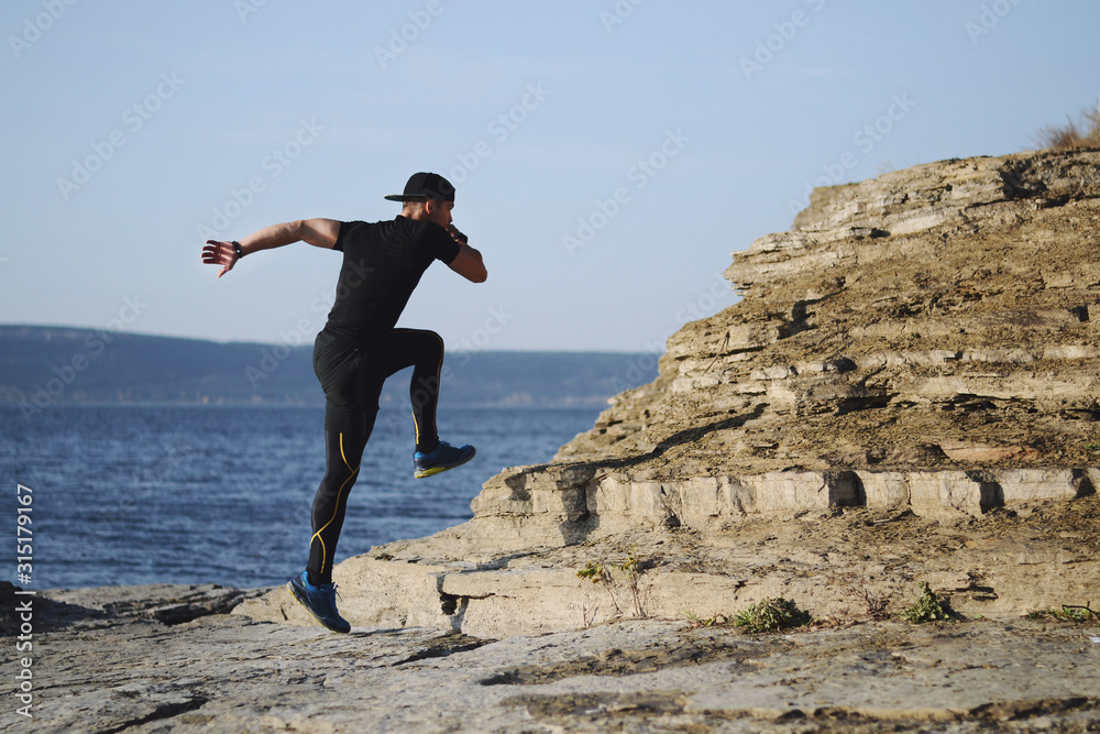 Male runner running on a mountain trail. Athlete runs in the mountains among the rocks. Man in black jersey and black shorts training outdoors Male runner running on a mountain trail. Athlete runs in the mountains among the rocks. Man in black jersey and black shorts training outdoors