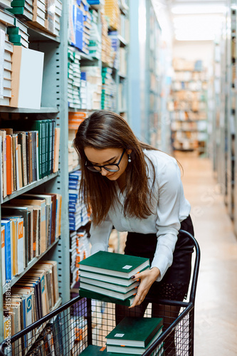 Young librarian worker with new books. Librarian puts piles of new books to the shelves of the library, doing her job