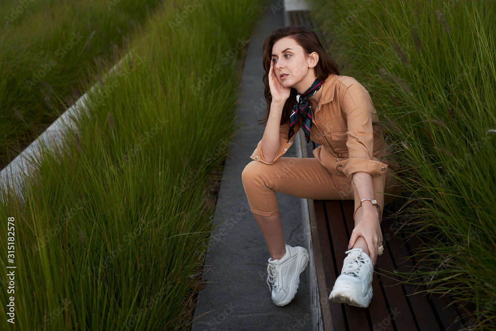 Young beautiful red haired gen z girl wearing slim fitted women beige khaki coverall overall denim jumpsuit or flight suit and white chunky platform sneakers sitting on a long park bench in high green