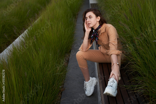 Obraz na płótnie Young beautiful red haired gen z girl wearing slim fitted women beige khaki coverall overall denim jumpsuit or flight suit and white chunky platform sneakers sitting on a long park bench in high green w salonie