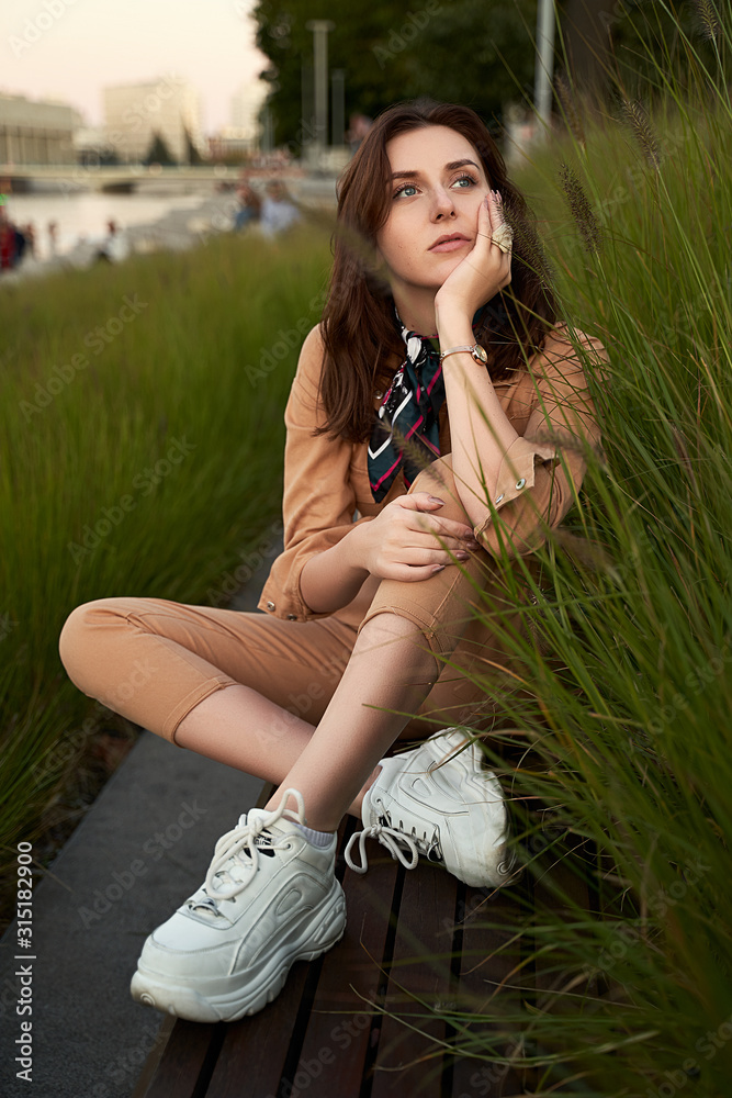 Young beautiful red haired gen z girl wearing slim fitted women beige khaki coverall overall denim jumpsuit or flight suit and white chunky platform sneakers sitting on a long park bench in high green