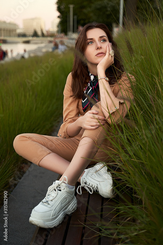 Obraz na płótnie Young beautiful red haired gen z girl wearing slim fitted women beige khaki coverall overall denim jumpsuit or flight suit and white chunky platform sneakers sitting on a long park bench in high green w salonie