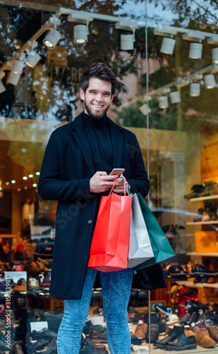 Young attractive bearded man with bags goes shopping in the city and looks at camera with his smartphone in hands.