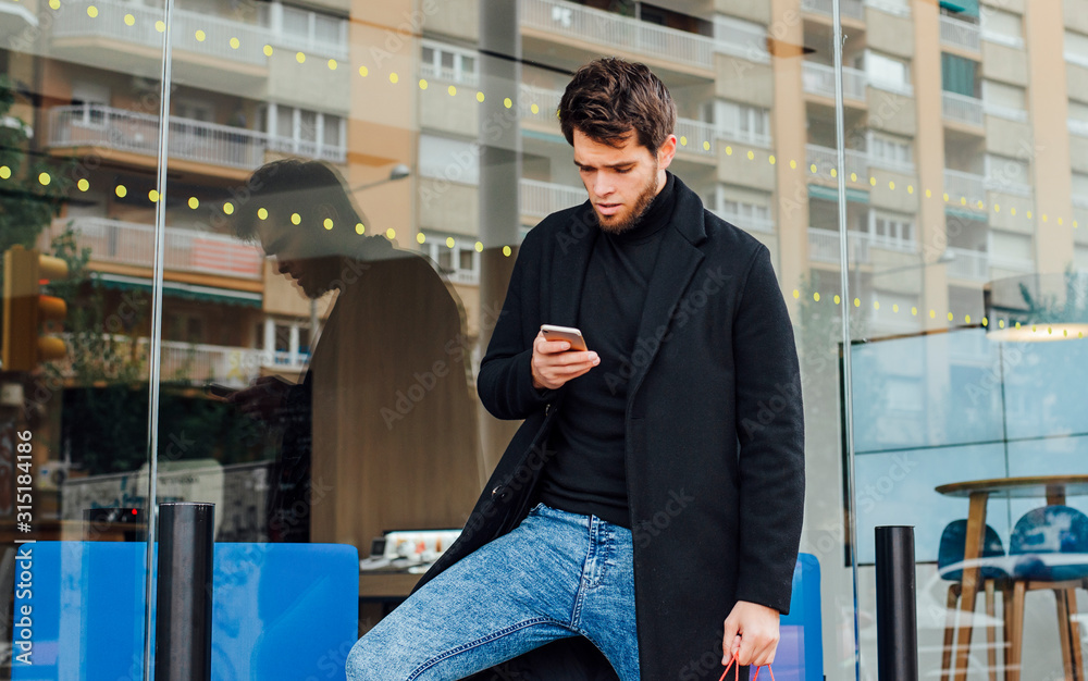 Fototapeta premium Attractive man with a beard and fashion with bags in his hands consults his smartphone in front of an office window.
