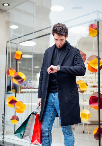 Young and attractive man holds bags in his hand waiting in front of a store and looks impatiently at his watch.