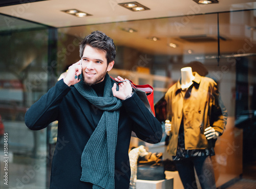 Attractive young man with a beard who has bags in one hand is talking on the phone while shopping in the city.