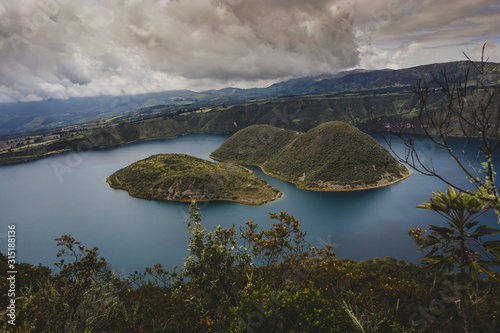 Looking towards the island on famous Lake Cuicocha in Ecuador. Panorama over a blue lake, surrounded with lush greens and white clouds.
