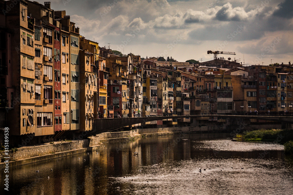 Dark looking panorama of Girona from the river, looking away from Girona cathedral of Saint Mary and red Eiffel bridge. Greenery on the water is seen.