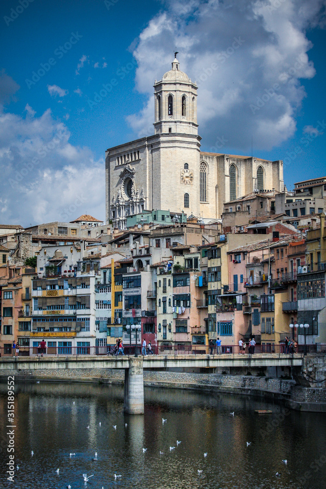 Panorama of Girona from the river, with Girona cathedral of Saint Mary and a bridge seen in the foreground. Some clouds on the sky behind the church.