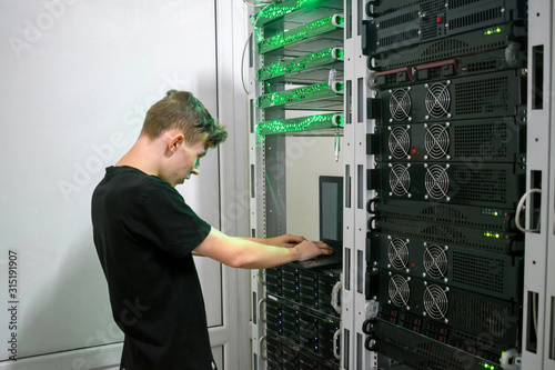 Young man works at a laptop near the racks with computer equipment. A teenager programmer programs server hardware in a modern data center. Information Technology Concept.
