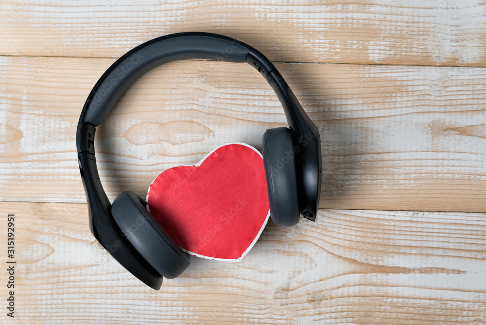 Full-size wireless headphones pulled over small red heart-shaped box on a light brown wooden table. Love music concept. Directly above Full-size wireless headphones pulled over small red heart-shaped box on a light brown wooden table. Love music concept. Directly above