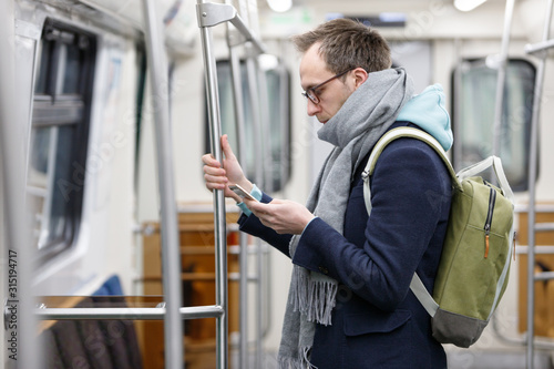 Caucasian young man passenger in eyeglasses with backpack holding handrail, using and looking at smart mobile phone at underground train. Subway lifestyle