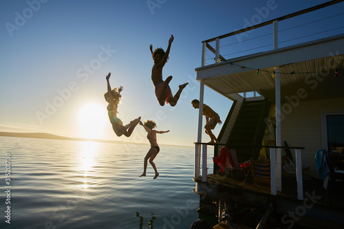 Young adult friends jumping off summer houseboat into sunset ocean