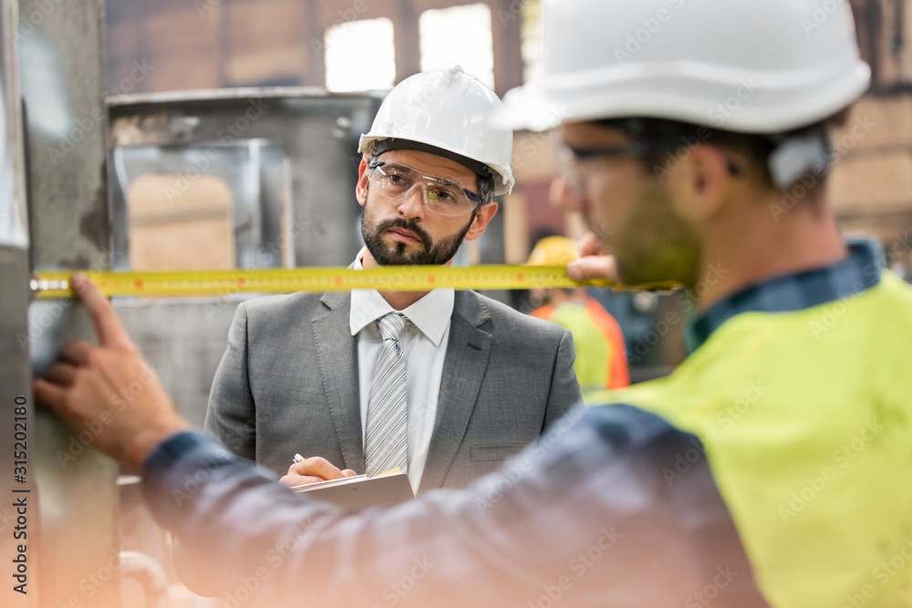 Manager watching steel worker with tape measure in factory Manager watching steel worker with tape measure in factory