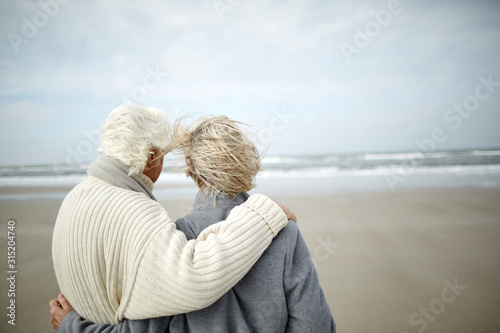 Pensive senior couple hugging looking at ocean view on windy winter beach photo