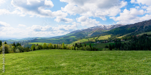mountainous countryside landscape in spring. grassy meadow on top of a hill. mountain ridge with snow capped tops in the distance. sunny weather with clouds on the blue sky © Pellinni