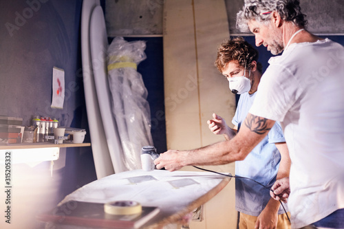 Male surfboard designers wearing protective masks sanding surfboard in workshop photo