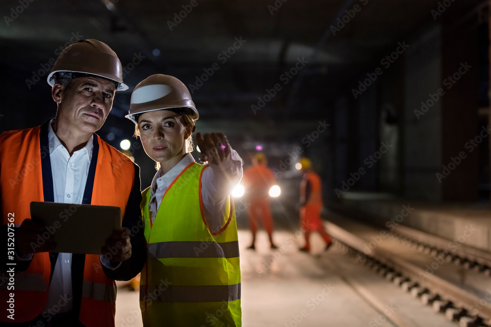 © Agnieszka Olek/Caia Image - Foreman engineer digital tablet talking on dark tracks at underground construction site