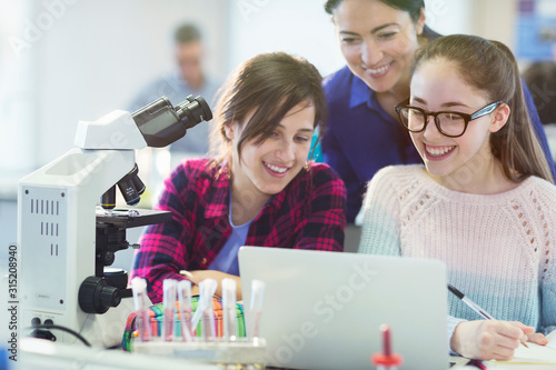 Female teacher and girl students using microscope and laptop, conducting scientific experiment in laboratory classroom