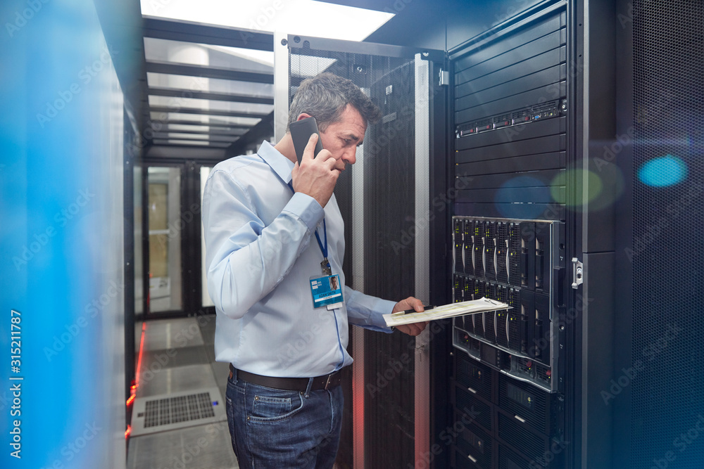 Male IT technician clipboard talking on cell phone at panel in server room Male IT technician clipboard talking on cell phone at panel in server room