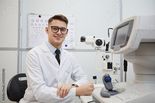 Portrait of male optometrist smiling at camera while posing at workplace by optic equipment, copy space