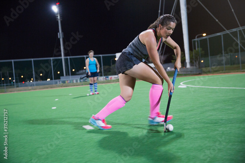 Focused young female field hockey player playing on field at night