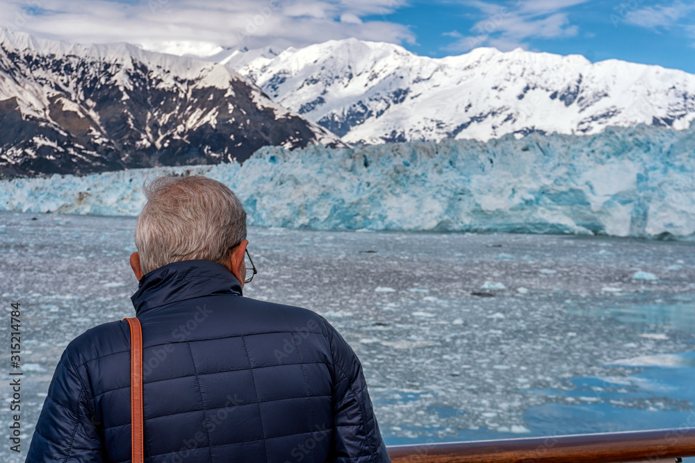 Old man is standing on the open deck of the cruise ship. Passenger is looking on the Hubbard Glacier in Alaska. Icebergs are around the boat. Amazing view on the mountains of the national park.
