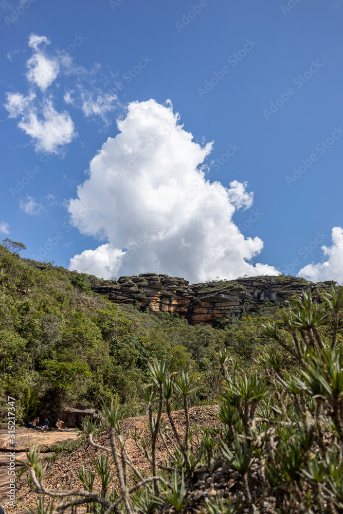 Huge plateau with sticking out rock formations in the Andorinhas [swallows] park in Ouro Preto seen from a lower viewpoint in the valley against a blue sky with clouds and vegetation in the foreground