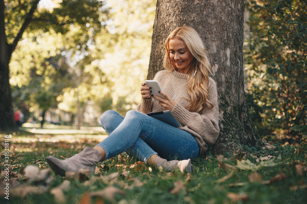 A woman sitting by a tree looking at her smartphone with headphones in her ears. A woman sitting by a tree looking at her smartphone with headphones in her ears.