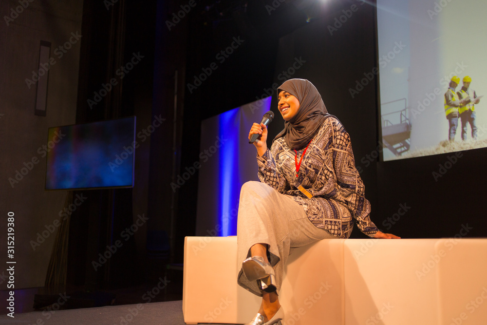 Smiling female speaker with microphone in hijab on stage Smiling female speaker with microphone in hijab on stage