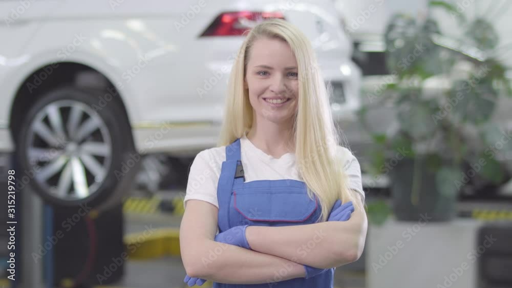 Portrait of young Caucasian woman standing at the background of white car in repair shop. Blond female auto mechanic crossing hands and smiling. Lifestyle, occupation, repair shop.