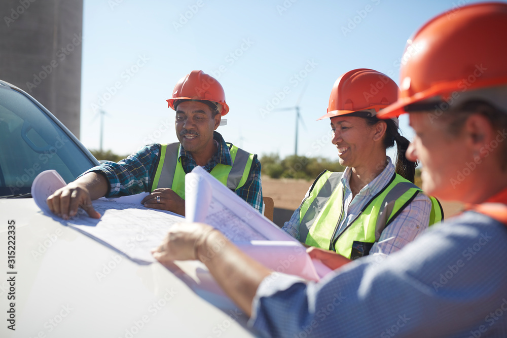 Engineers reviewing blueprints on truck at sunny wind turbine power plant Engineers reviewing blueprints on truck at sunny wind turbine power plant