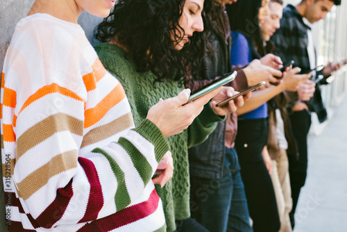 Slika na platnu Young men and women look at their smart phones - Standing against a wall in the