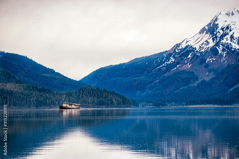 Fishing boat is sailing between the snow peak mountains and Tongass National forest. Beautiful reflection in the water. Vessel is looking for a salmon. 