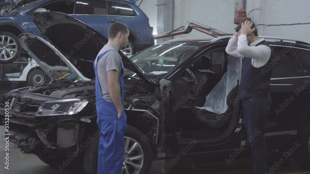Side view of shocked Caucasian man standing next to his broken car as auto mechanic coming up. Desperate car owner driving automobile for repair. Car accident, repair shop.