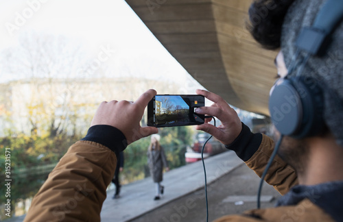 Young man with headphones using camera phone along canal