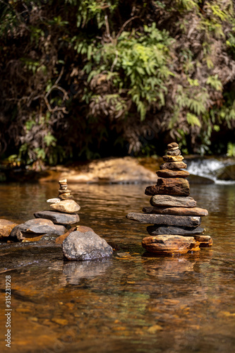 Small rocks stacked on top of each other in a pond part of a stream leading to a waterfall with a small cascade out of focus in the background