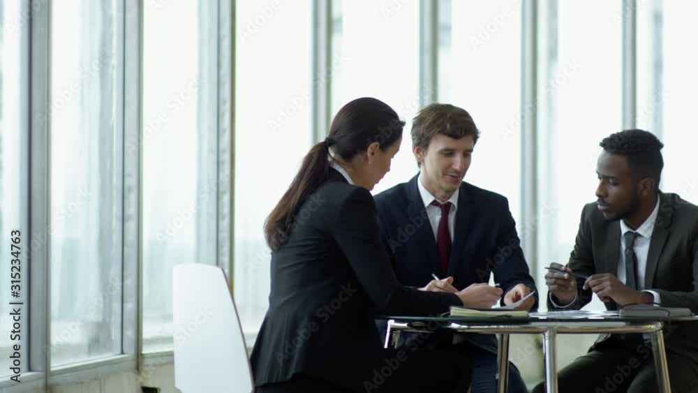 PAN of businesswoman and businessmen in suits sitting at table in conference room with panoramic windows and discussing documents PAN of businesswoman and businessmen in suits sitting at table in conference room with panoramic windows and discussing documents