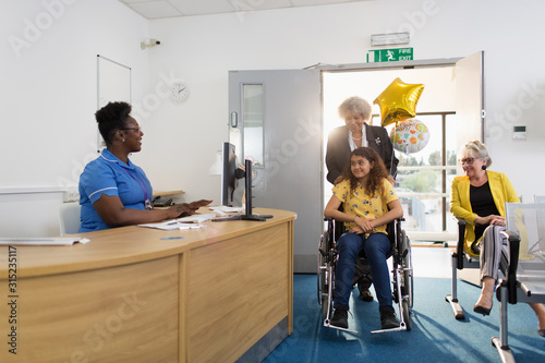 Woman pushing girl patient in wheelchair into clinic reception photo