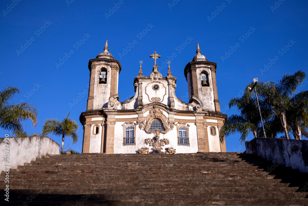 Stairs in the foreground leading up to Our Lady of Carmo church in historic colonial city centre of Ouro Preto with palm trees to the side against a clear blue sky