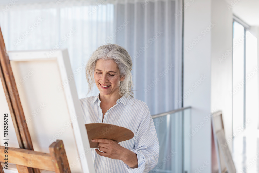 Smiling mature woman with palette painting at canvas on easel