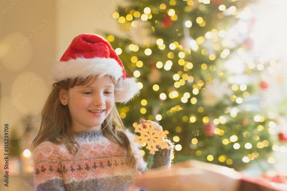Girl in Santa hat holding snowflake ornament in front of Christmas tree Girl in Santa hat holding snowflake ornament in front of Christmas tree