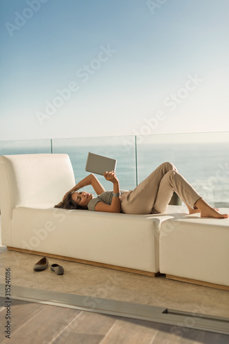Woman using digital tablet relaxing laying on chaise lounge on luxury balcony with ocean view